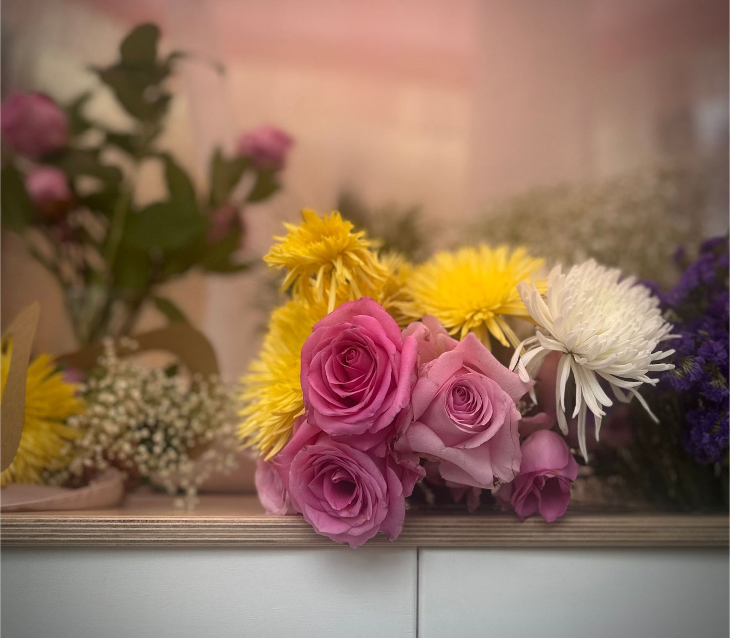 Bouquet of pink roses and yellow chrysanthemums on a wooden surface with a blurred floral background.